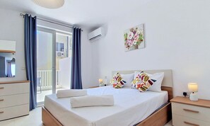 A hotel room with a bed, a nightstand, a lamp, a wall-mounted air conditioner, a window with curtains, and a balcony. - Modern 2BR Flat · Balcony · St Paul's Bay (San Pawl il-Baħar)