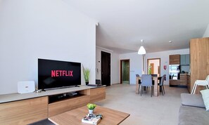Ein modernes Wohnzimmer mit einem Flachbildfernseher, der 'NETFLIX' zeigt, einem Essbereich mit Tisch und Stühlen sowie einer Küche im Hintergrund.