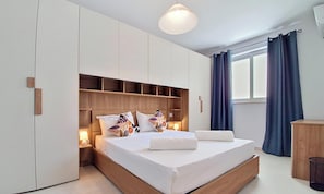 A modern bedroom with a wooden headboard, a bed with white linens, a nightstand with a lamp, and a window with curtains. - Modern 3BR Flat · Balcony · St Paul's Bay (San Pawl il-Baħar)