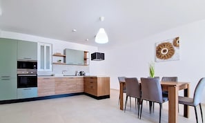 A modern kitchen with built-in appliances, a dining area with a wooden table and chairs, and a wall-mounted light fixture. - Modern 3BR Flat · Balcony · St Paul's Bay (San Pawl il-Baħar)