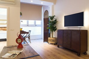 Una habitación con piso de madera, un reloj rojo, una mesa con un periódico, una planta, un armario y un televisor en la pared.
