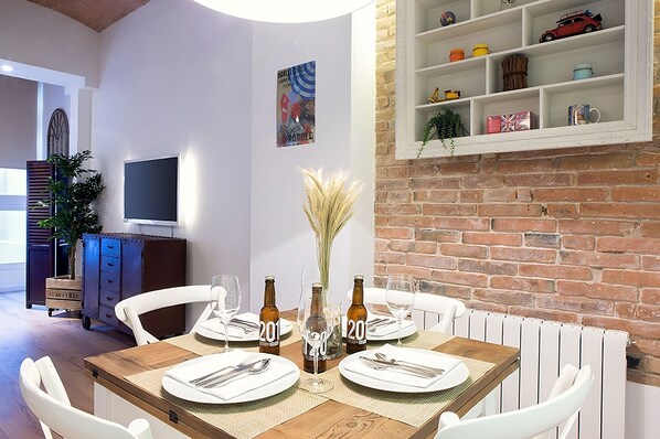 Un comedor con pared de ladrillo, estanterías blancas con adornos, una mesa de madera preparada para cuatro personas y un televisor en la pared.