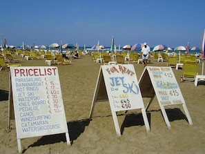 Beachside chairs, umbrellas, and signs advertising water sports and rental prices. - Gouves Sunset Apts (Kato Gouves)