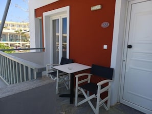 A balcony with a white table and two chairs, a white door, and a red wall with a number plaque. - Gouves Sunset Apts (Kato Gouves)
