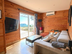 A wooden interior with a sofa, a table, a TV, and a sliding glass door leading to an outdoor area. - Wooden house / Swimming pool / Private Guinguette / Sea view / Air conditioning / Jacuzzi optional (Le Beausset)