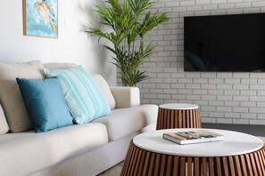 A living room with a sofa, pillows, a round wooden side table, a book, a potted plant, and a TV mounted on a brick wall. - The Turtle's Nest in Safety Bay - Near the Beach (Safety Bay)
