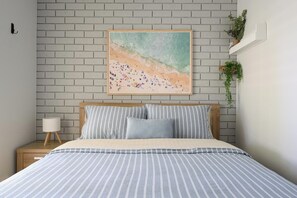 A bedroom with a bed, a nightstand, a lamp, a brick wall, and a painting of a beach scene. - The Turtle's Nest in Safety Bay - Near the Beach (Safety Bay)