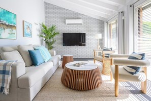 A modern living room with a white brick wall, a flat-screen TV, a beige sofa, a round wooden coffee table, and a wicker chair. - The Turtle's Nest in Safety Bay - Near the Beach (Safety Bay)