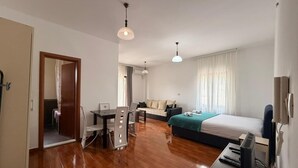 A modern hotel room with a wooden floor, a bed with a green blanket, a desk with a computer, a chair, and a TV. - Apartments Oasis (Markovići)
