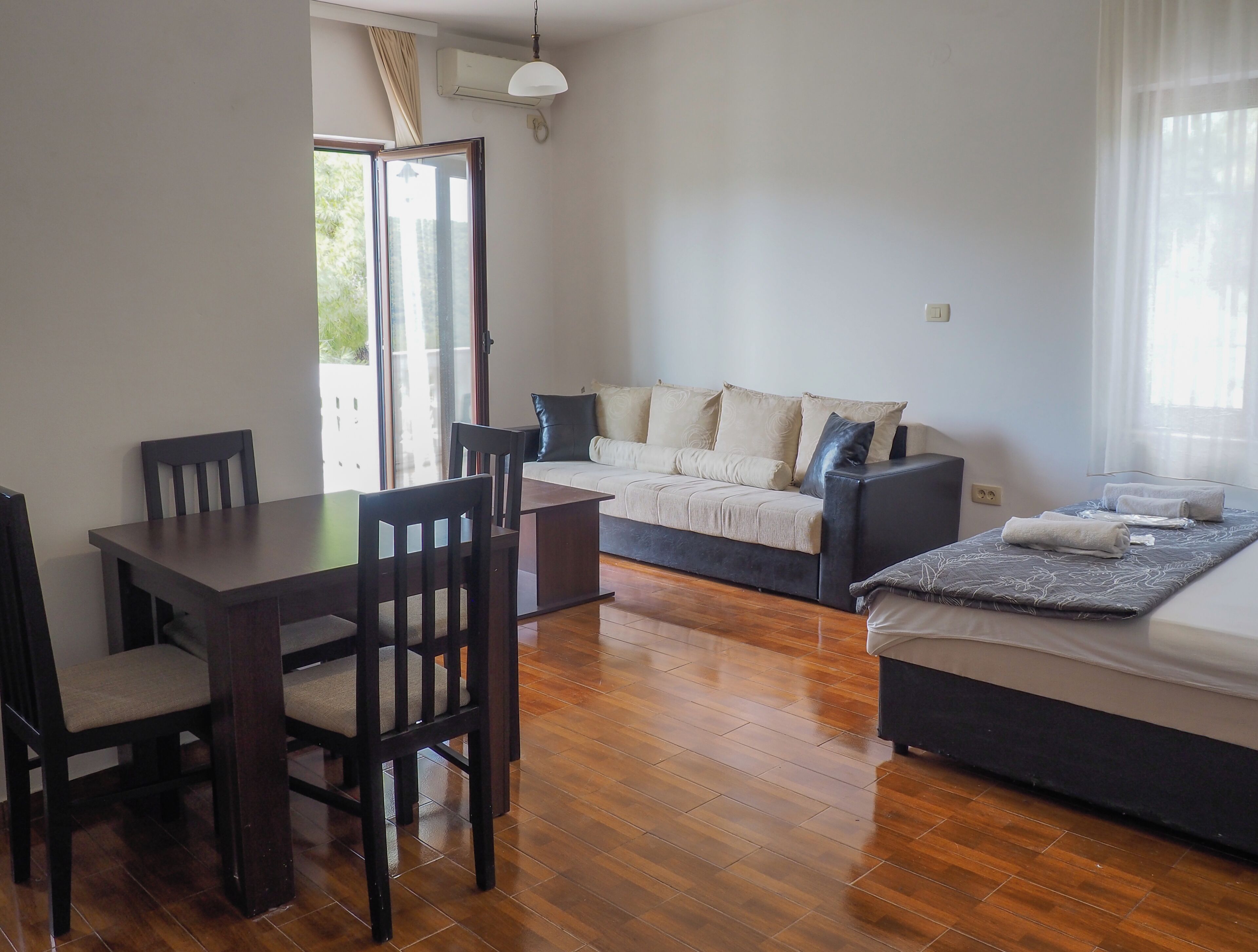 A room with a wooden dining table, two chairs, a sofa, a bed with a blanket, and a window with curtains.