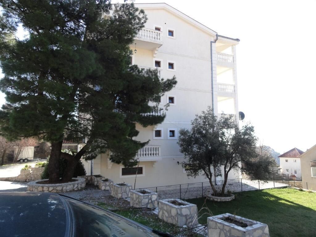 A multi-story building with balconies, a parking area, and a tree in the foreground.