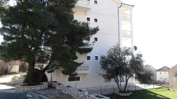 A multi-story building with balconies, a parking area, and a tree in the foreground.