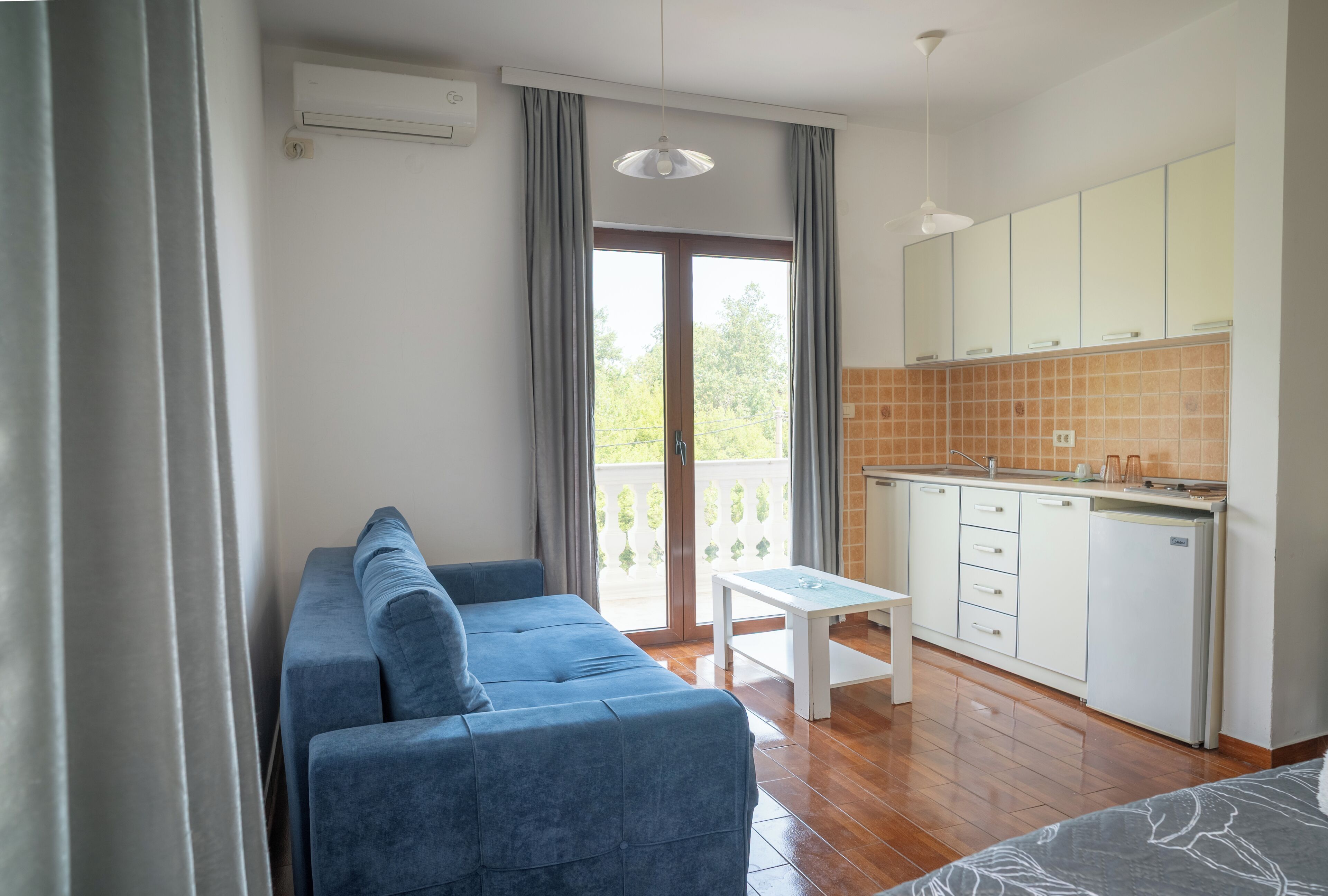 A room with a sofa, a small table, a kitchen area, and a sliding glass door leading to an outdoor balcony.