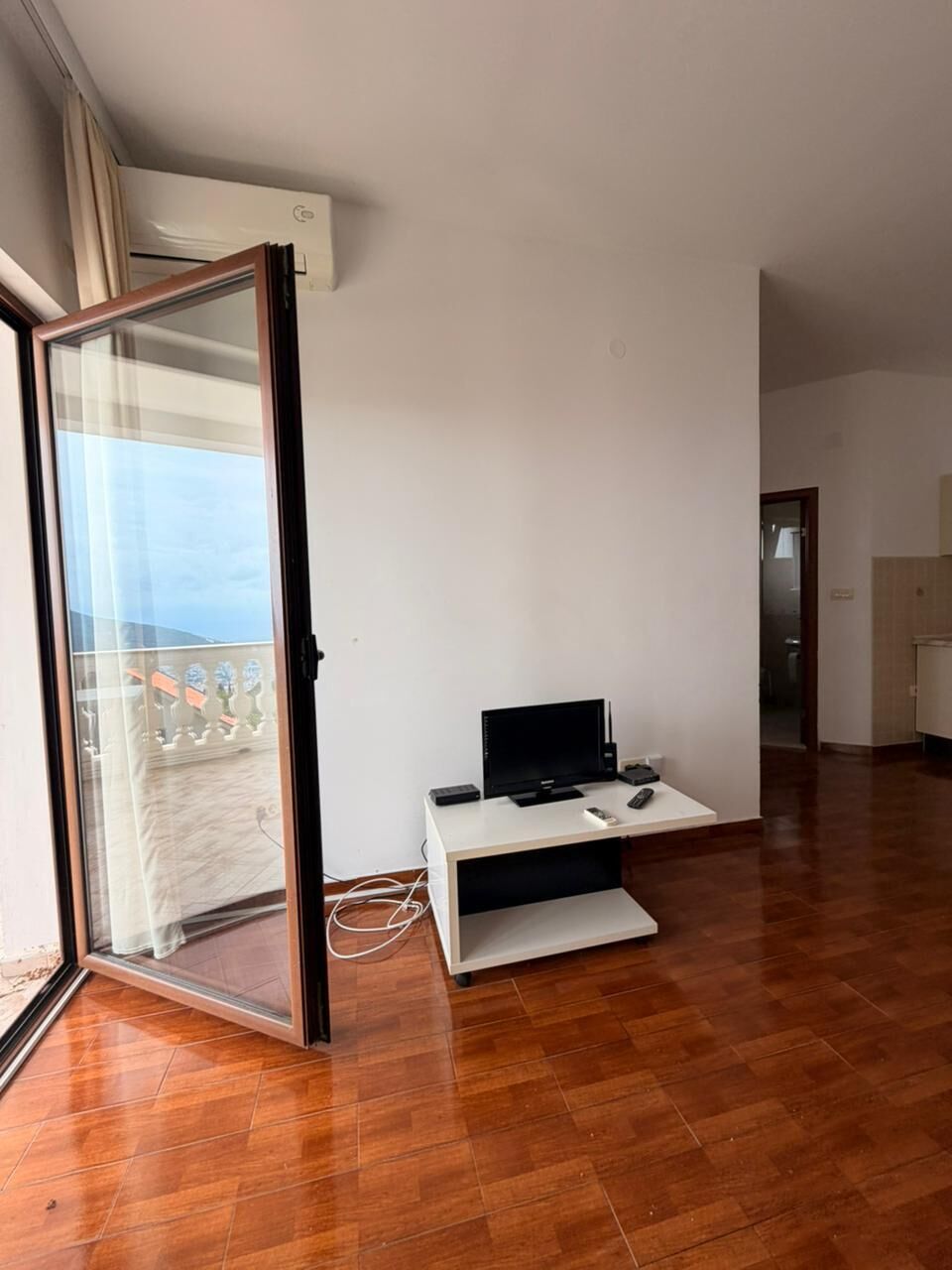 Una habitación con puerta de vidrio corredera que da a un balcón, un escritorio blanco con computadora y controles remotos, y piso de madera.
