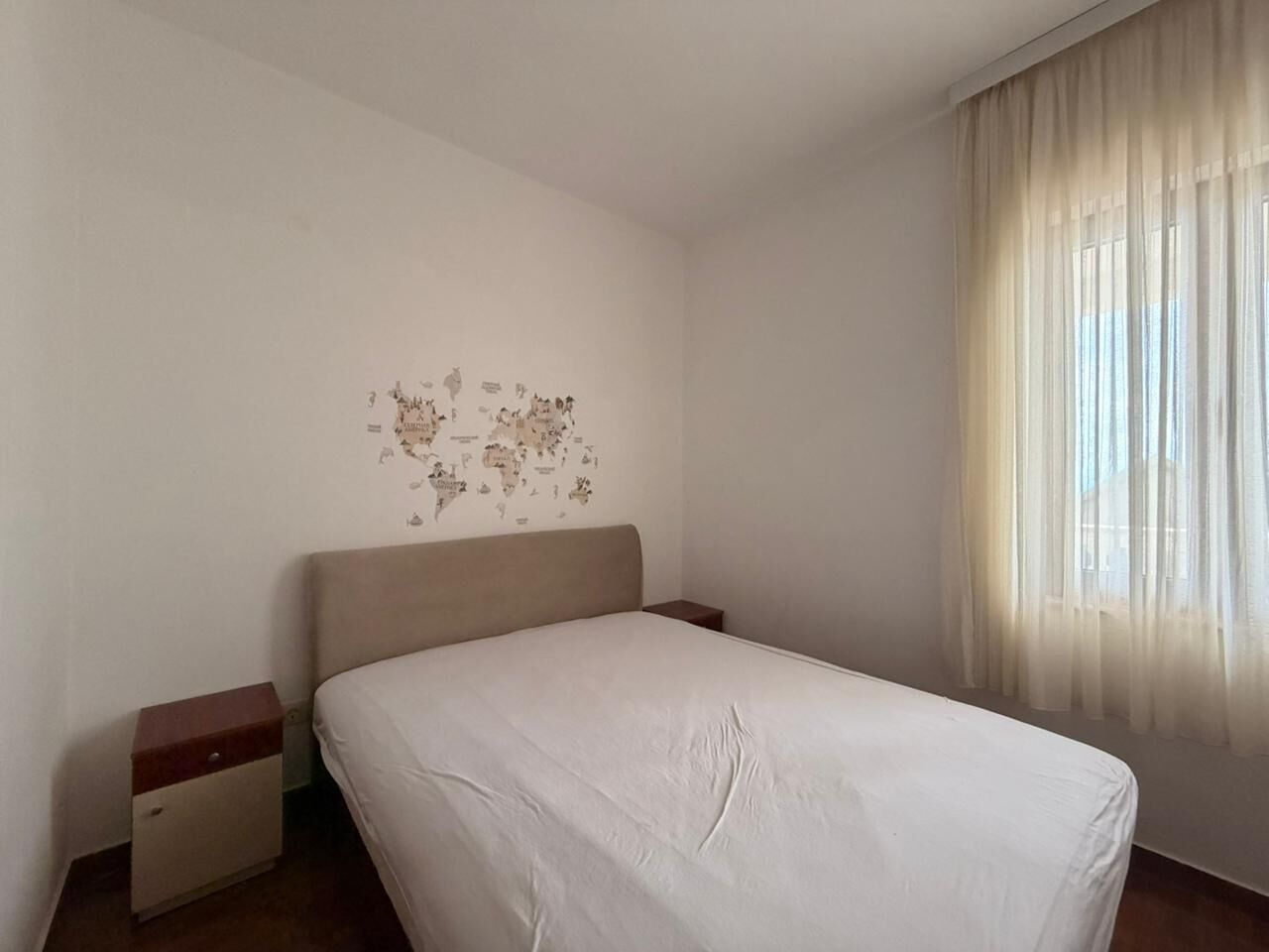 A hotel room with a bed, nightstands, a world map wall decoration, and a window with sheer curtains.