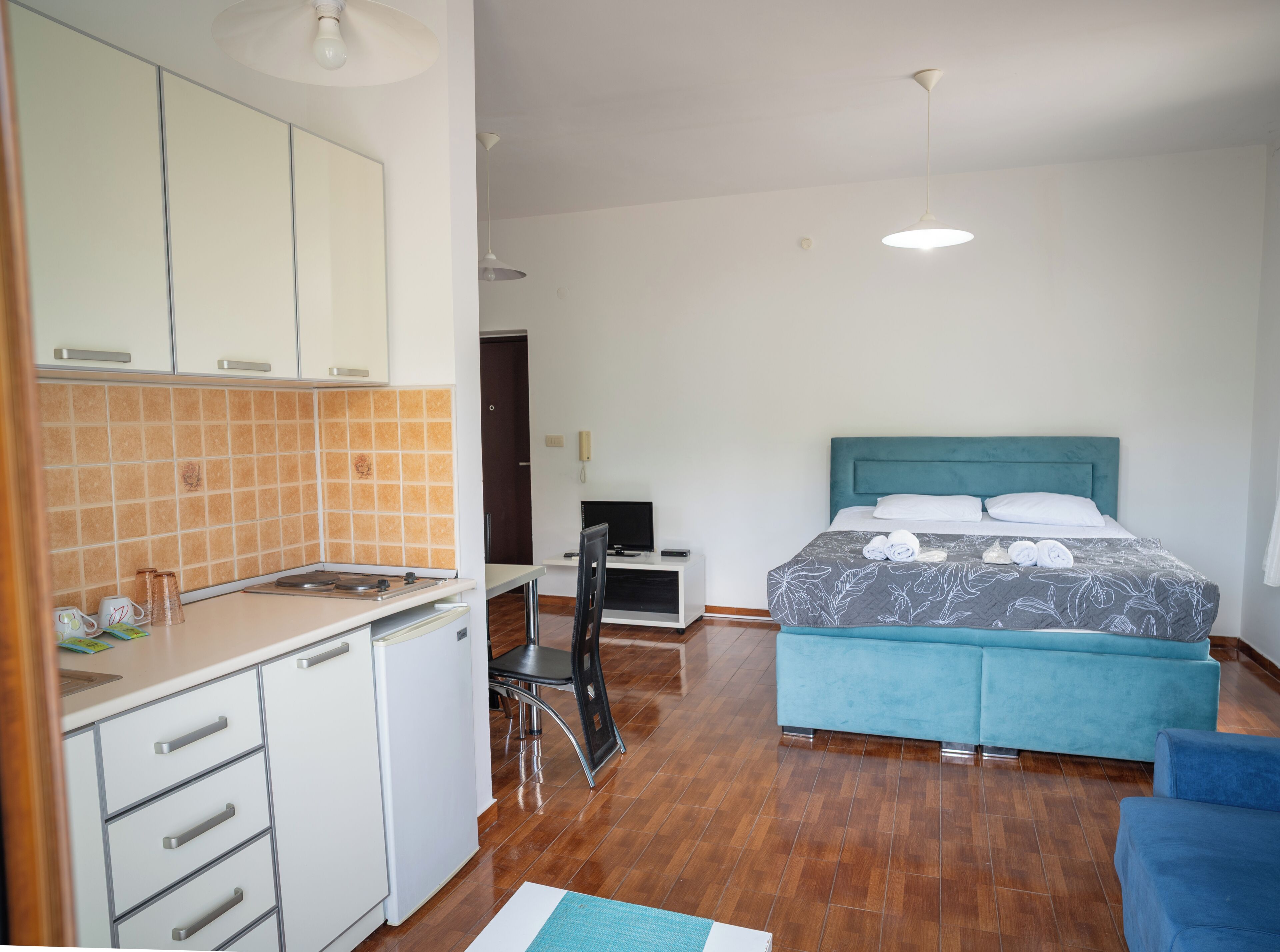 Habitación de hotel compacta con kitchenette, una cama con cabecera azul y un pequeño escritorio con silla.