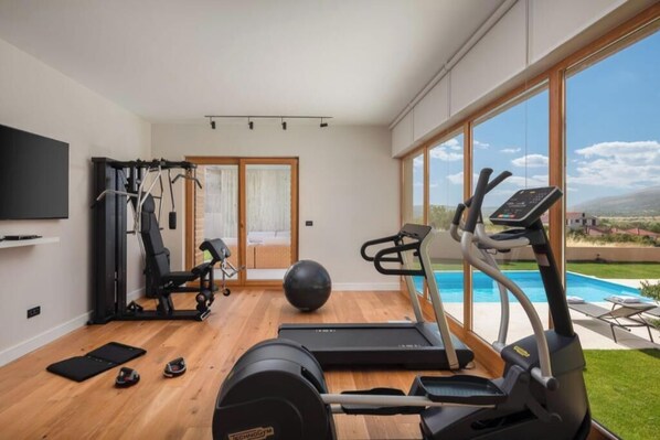 Een sportschool in huis met trainingsapparatuur, een televisie en uitzicht op een zwembad.