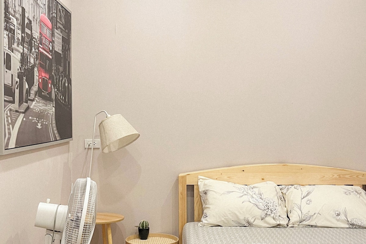 Un dormitorio con cama, un ventilador, una mesita con una planta, una lámpara y un cartel en la pared.