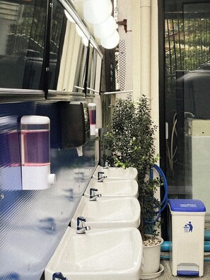 A row of sinks with faucets, a potted plant, and a trash bin with a recycling symbol. - Basecamp Hostel (Chiang Mai)
