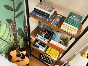 A bookshelf with various books, a guitar, and a checkered board game. - Basecamp Hostel (Chiang Mai)
