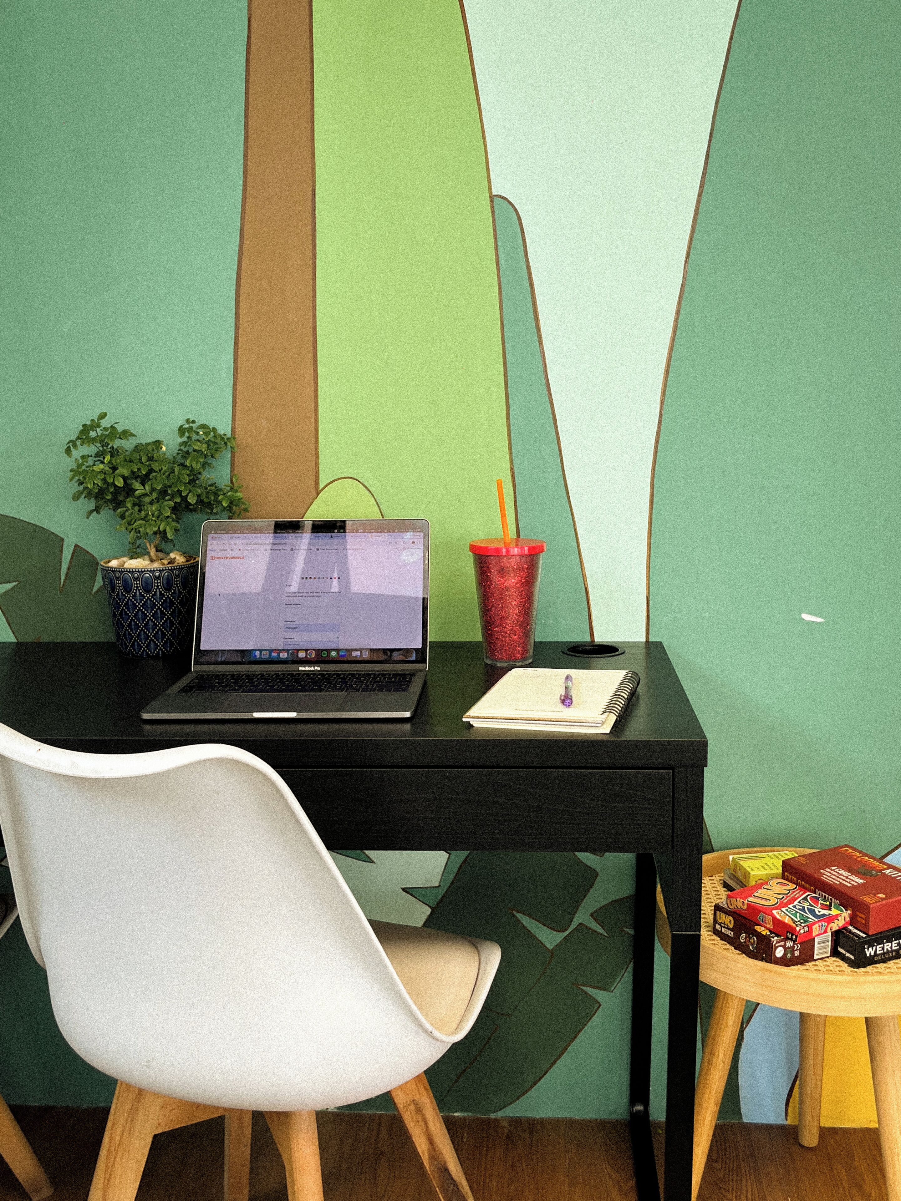 Ein Arbeitsplatz mit einem Laptop, einem Topf mit einer Pflanze, einer roten Tasse mit Trinkhalm, einem Notizbuch und einem kleinen Tisch mit Büchern.