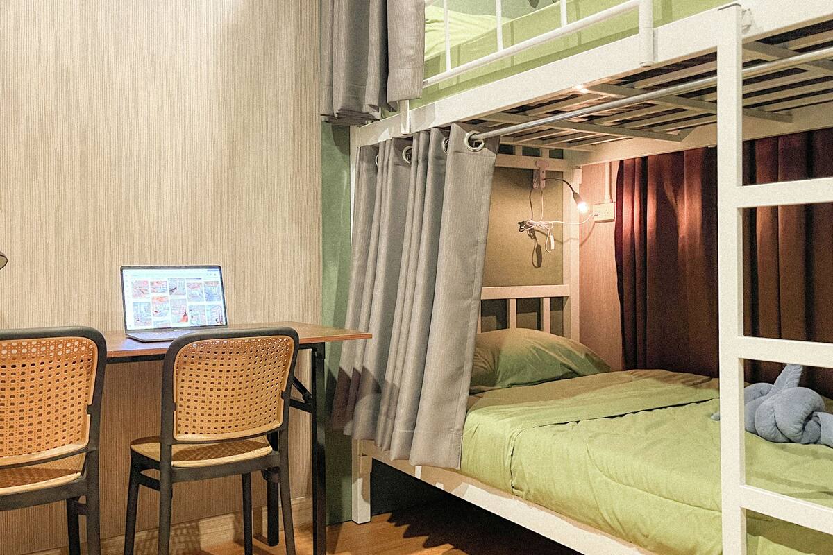 Una cama individual sobre otra con escritorio, sillas, una computadora portátil y una cortina.