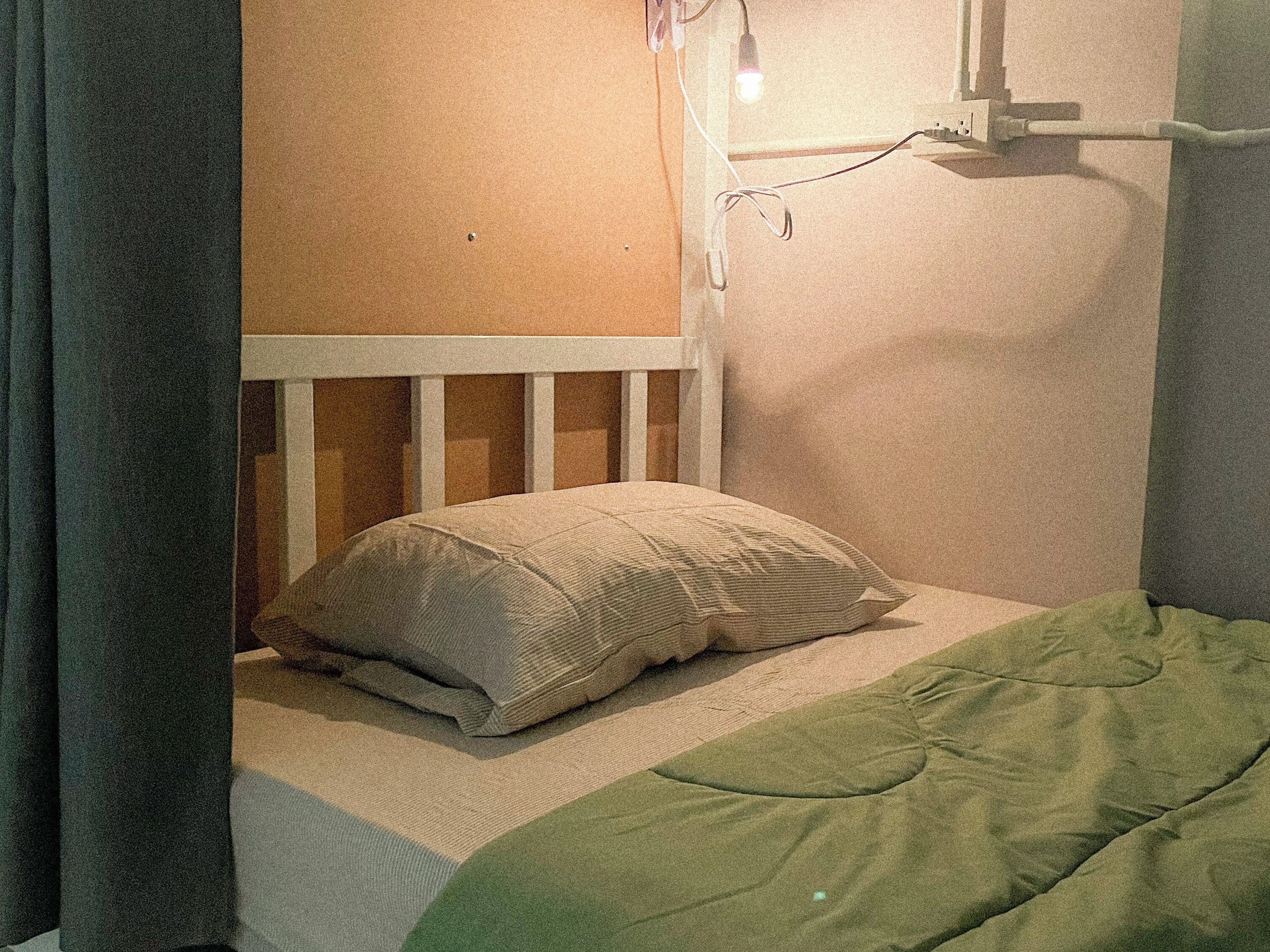 A single bed with a green blanket and a pillow in a room with a white headboard and a wall socket.