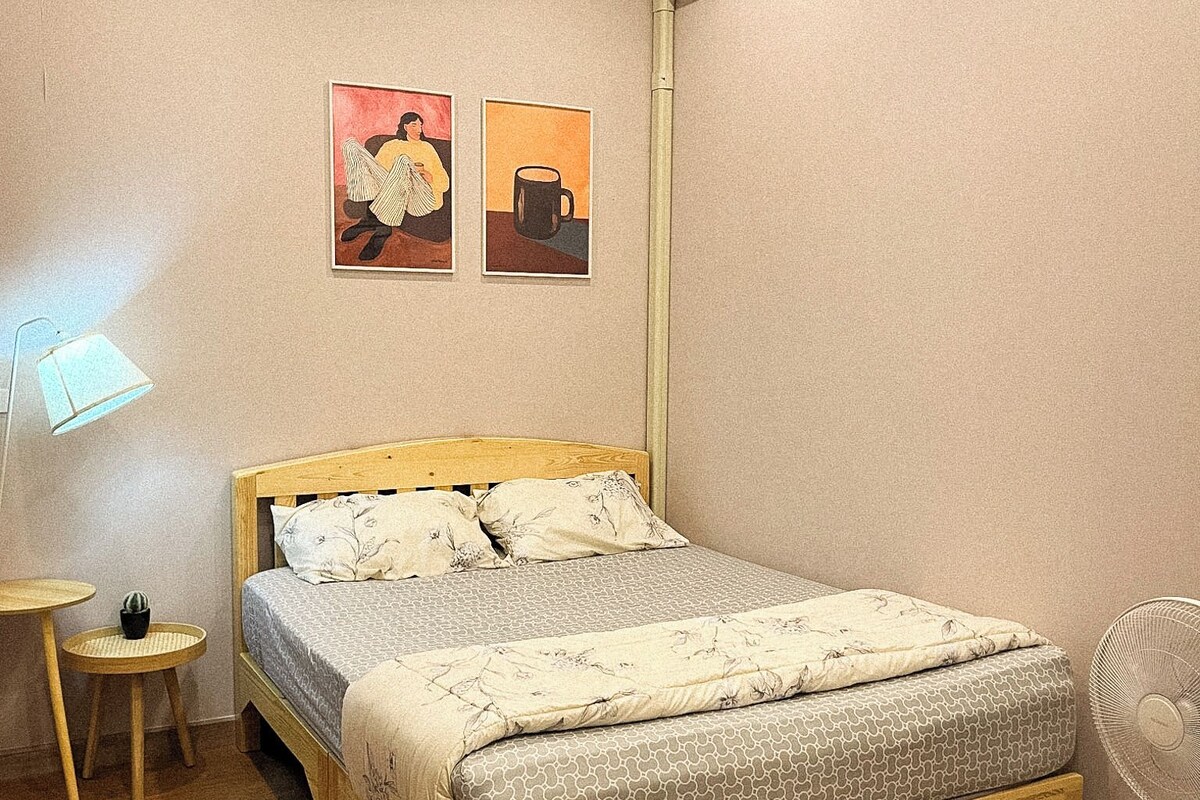 A bedroom with a wooden bed, a bedside table with a lamp, a small potted plant, and a wall-mounted air conditioner.