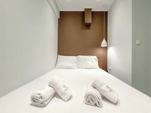 Une chambre d’hôtel avec un lit, deux serviettes enroulées, une lampe fixée au mur et une bouche d’aération au plafond.