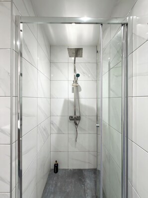 Een douche met een handdouche en een vaste douchekop, een fles shampoo, en een grijze betegelde vloer.