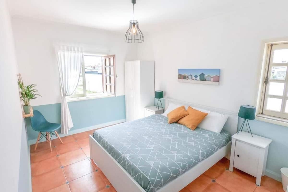 Un dormitorio con cama, una silla, una mesita de noche, una lámpara, una ventana y un cuadro en la pared.