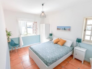 A bedroom with a bed, a chair, a nightstand, a lamp, a window, and a painting on the wall. - OLA Guesthouse (ESMORIZ)