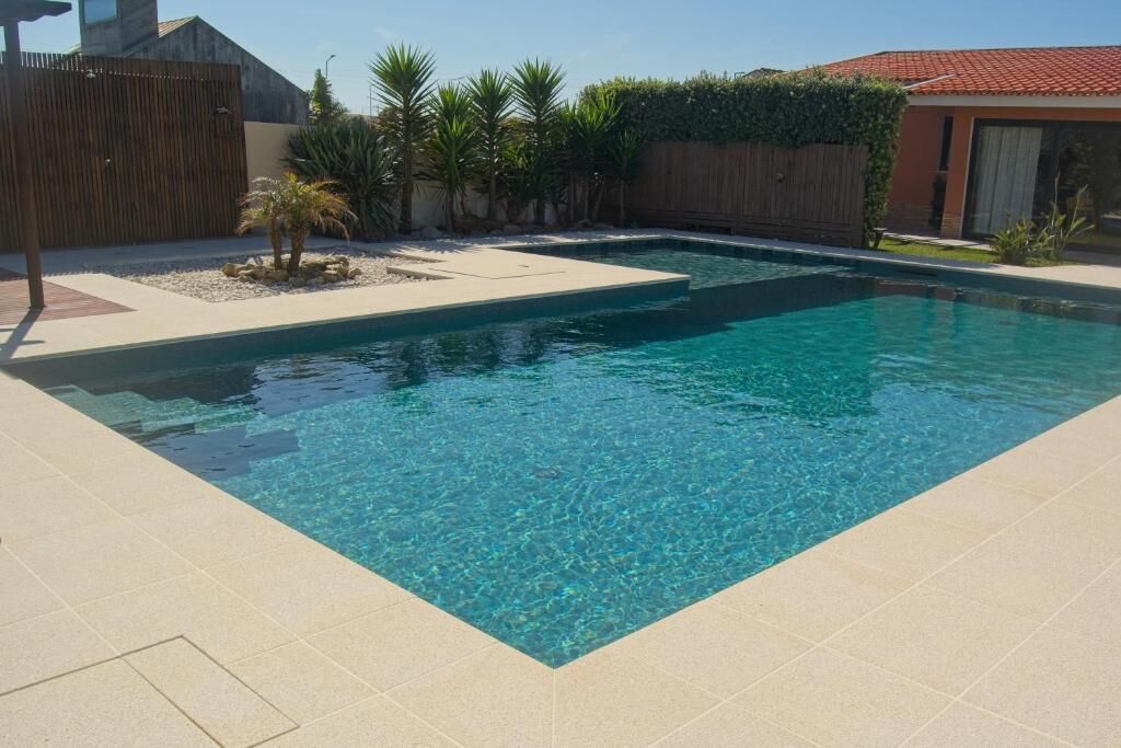 Una piscina rectangular con agua azul cristalina, rodeada por un suelo de baldosas y una valla de madera.
