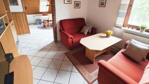 A cozy living room with a red sofa, a wooden coffee table, and a small table with a flower vase. There is a brick archway leading to a dining area with a wooden table and chairs. - Vacation apartment on the North Sea - Holtgast (East Frisia) (Holtgast)