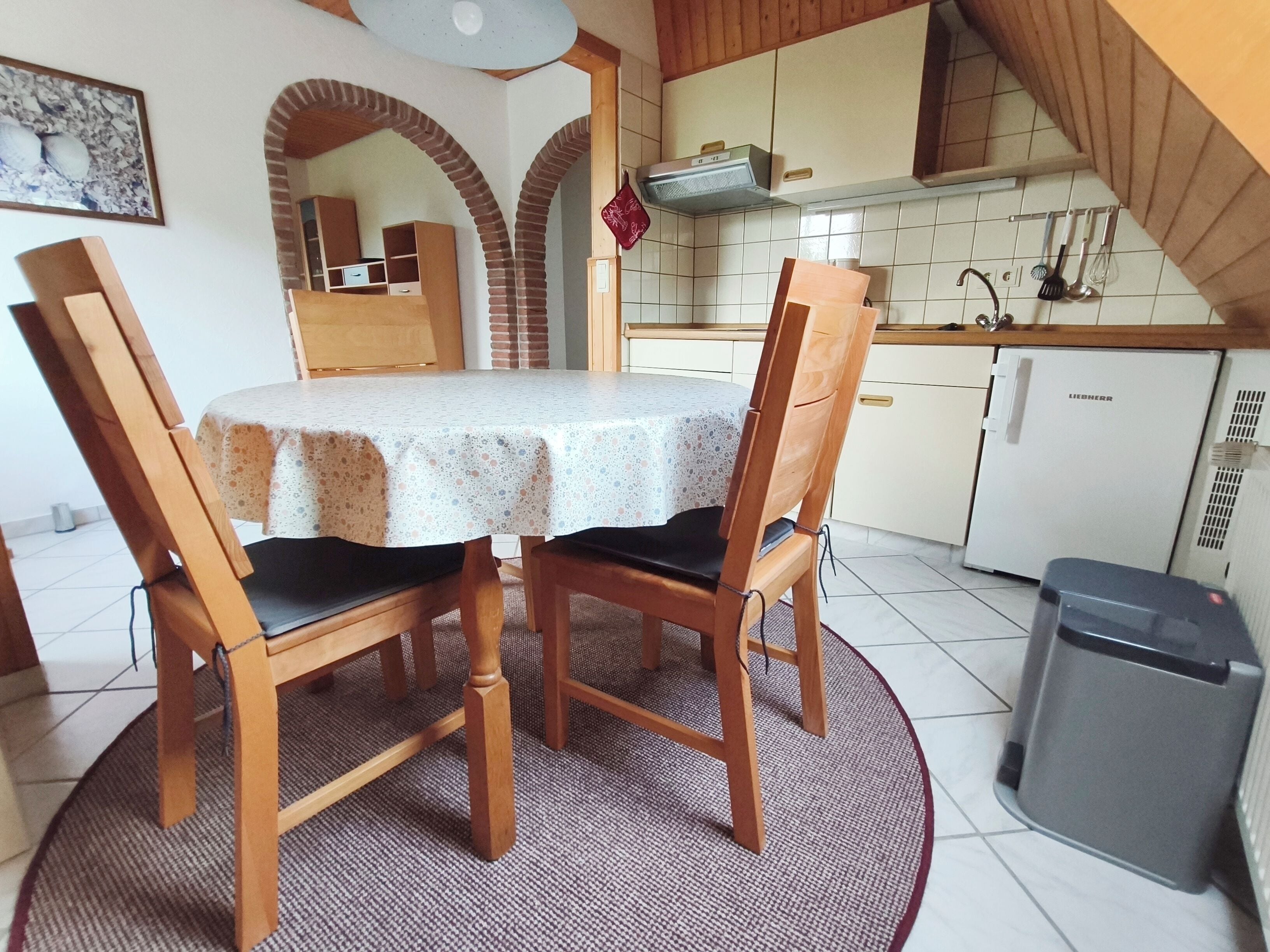 Un'area pranzo con un tavolo rotondo, sedie di legno, una tovaglia fantasia e una cucina sullo sfondo, più un cestino.