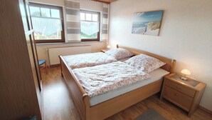 A bedroom with a wooden bed, two bedside tables, a radiator, a window with blinds, and a painting on the wall. - Vacation apartment on the North Sea - Holtgast (East Frisia) (Holtgast)