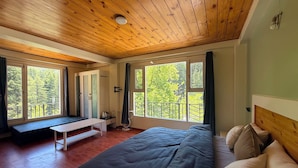 A bedroom with a wooden ceiling, large windows, a bed, a bench, and a small table. - Revivo The Confluenz (Manali)