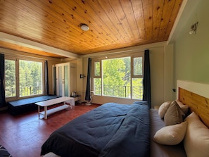 A bedroom with a wooden ceiling, large windows, a bed, a bench, and a small table. - Revivo The Confluenz (Manali)