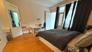 A bedroom with a large bed, a small desk, a chair, a radiator, and a window with curtains. - Revivo The Confluenz (Manali)