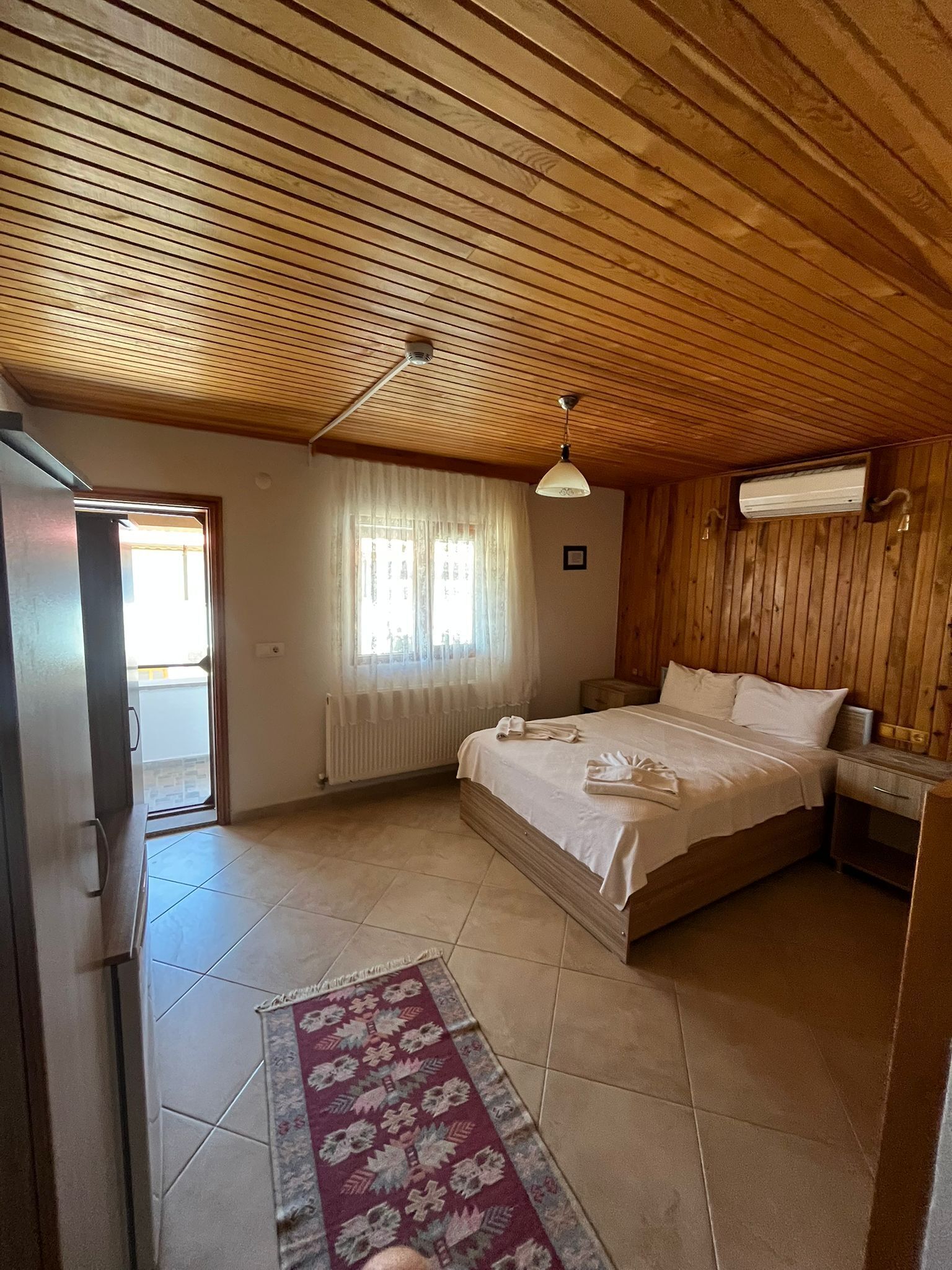A bedroom with a wooden ceiling, a bed with white linens, a nightstand, a window with curtains, and a radiator.