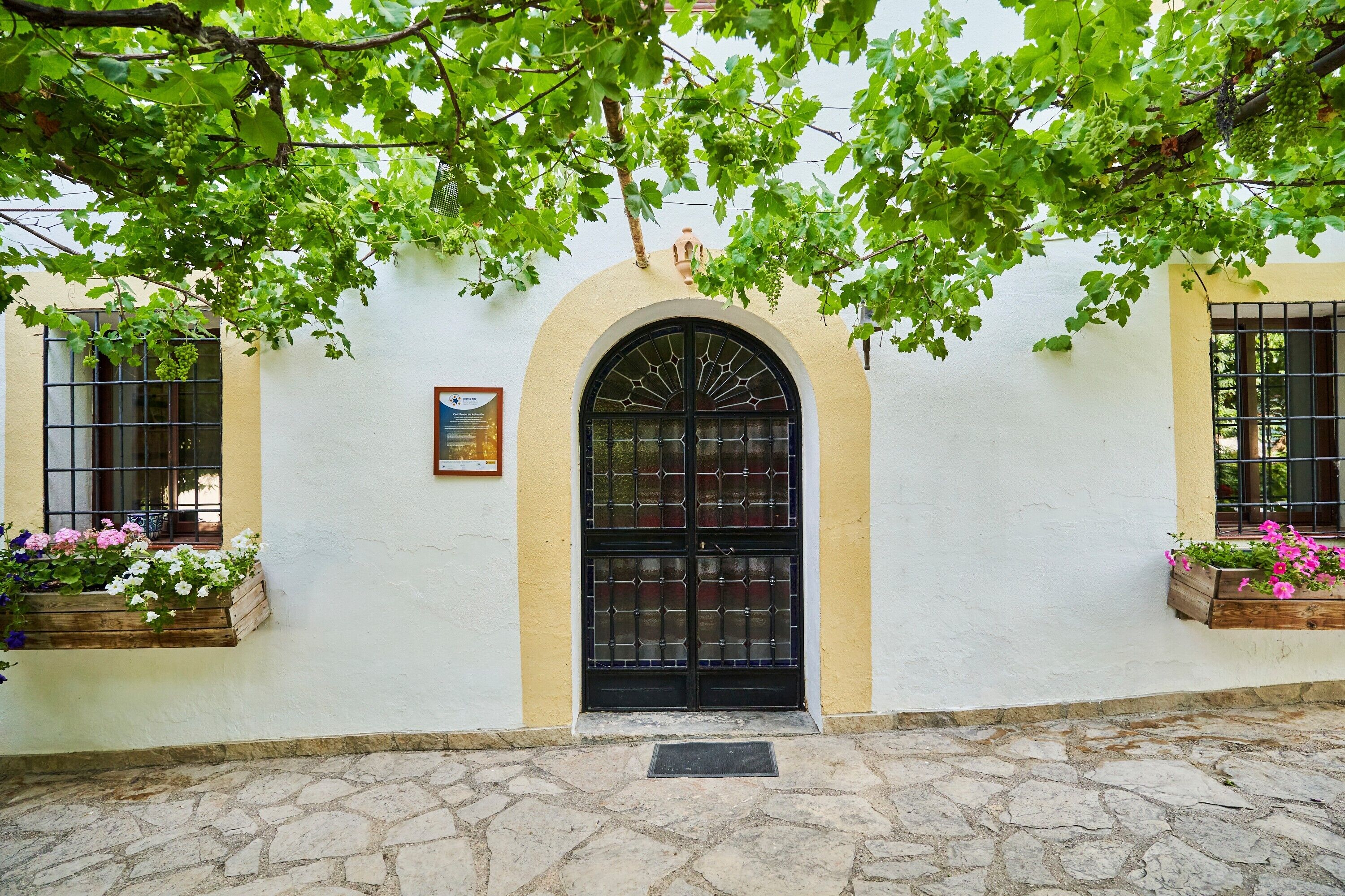 Un edificio con entrada empedrada de piedra, una puerta arqueada negra y macetas con flores rosadas.