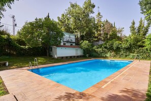 En rektangulär swimmingpool med en kakelbeklädd poolkant, omgiven av frodig grönska och en vit byggnad med ett rött tak.