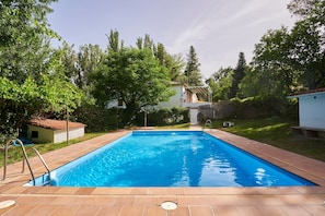 Una piscina rectangular con solárium de baldosas, rodeada de vegetación y un edificio pequeño al fondo.
