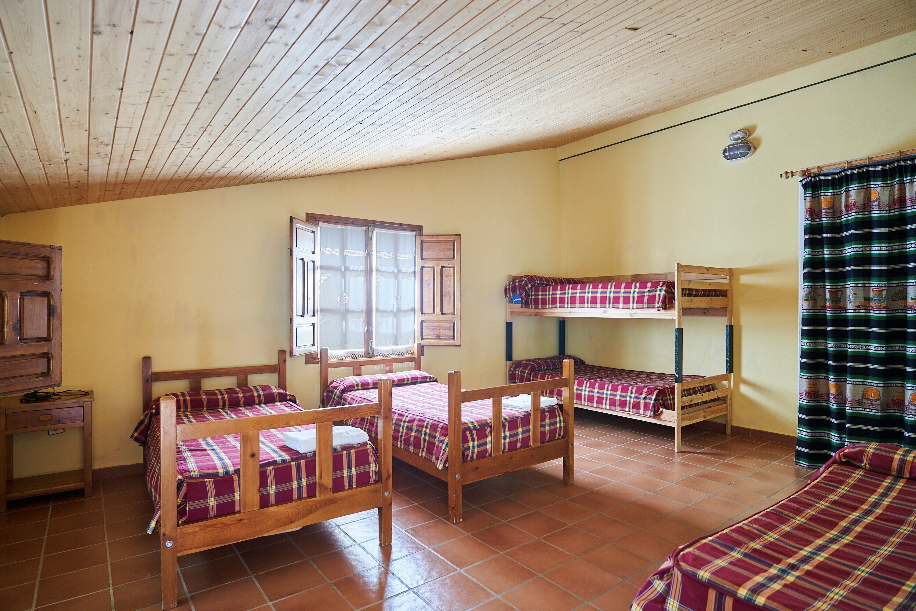 A dormitory room with bunk beds, a wooden desk, and a window with curtains.