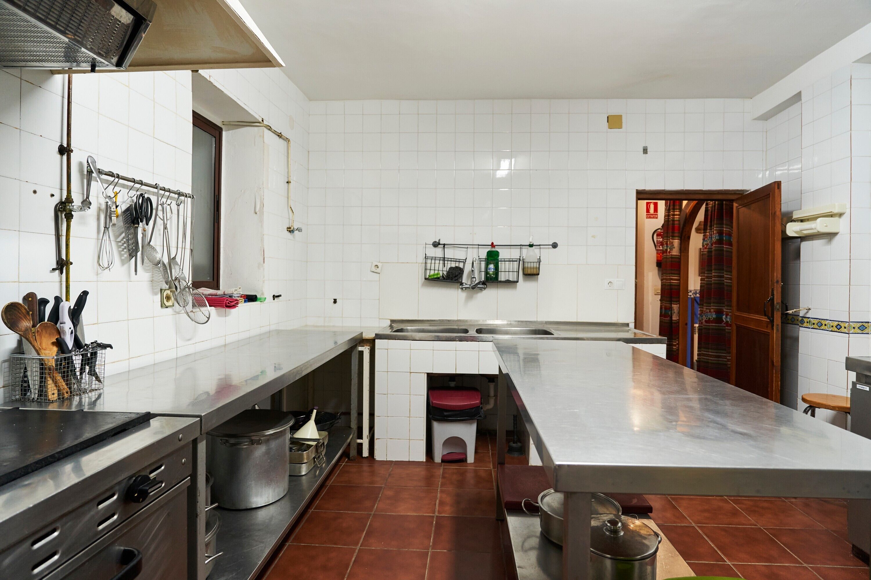 Una cocina con azulejos blancos, encimeras de acero inoxidable y diversos utensilios y electrodomésticos.