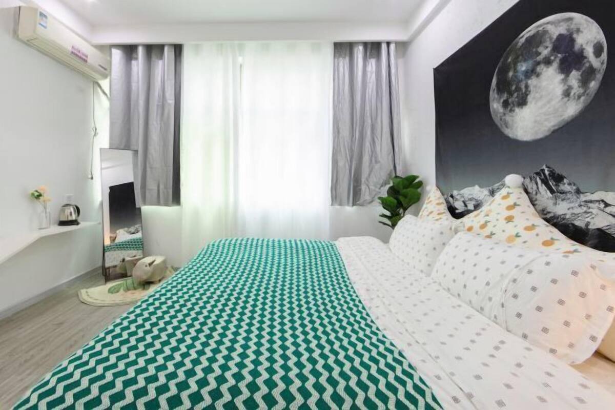 A bedroom with a bed, a painting of a moon, a window with curtains, and a plant.