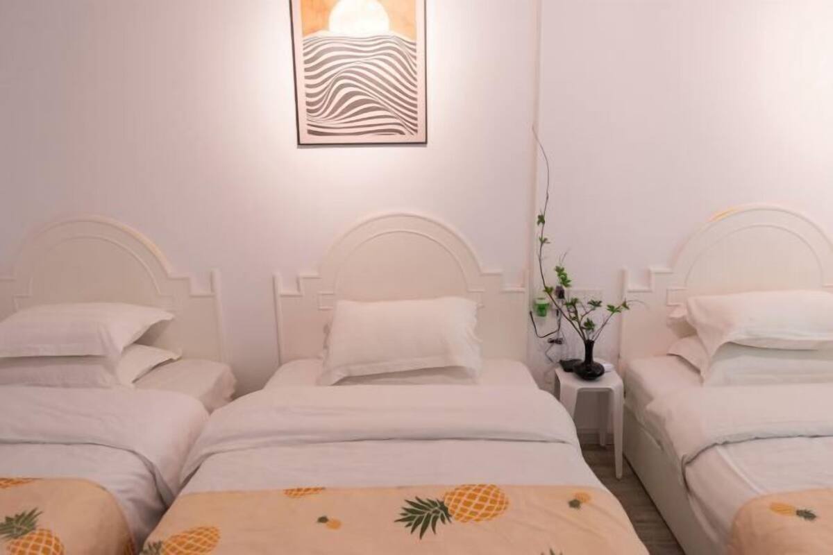 A room with three beds, each with a white headboard and white bedding. There is a wall-mounted artwork above the beds and a small table with a plant between them.