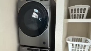 Ein Waschraum mit zwei übereinander gestapelten Waschmaschinen mit Frontlader, einem weißen Wäschekorb auf einer Ablage und einem Karton mit der Aufschrift 'HELLO FRESH' auf dem Boden.