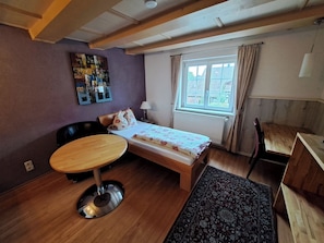 Une chambre à coucher avec un lit, une table ronde, une chaise, une lampe, une fenêtre avec des rideaux et un mur orné d’un tableau.