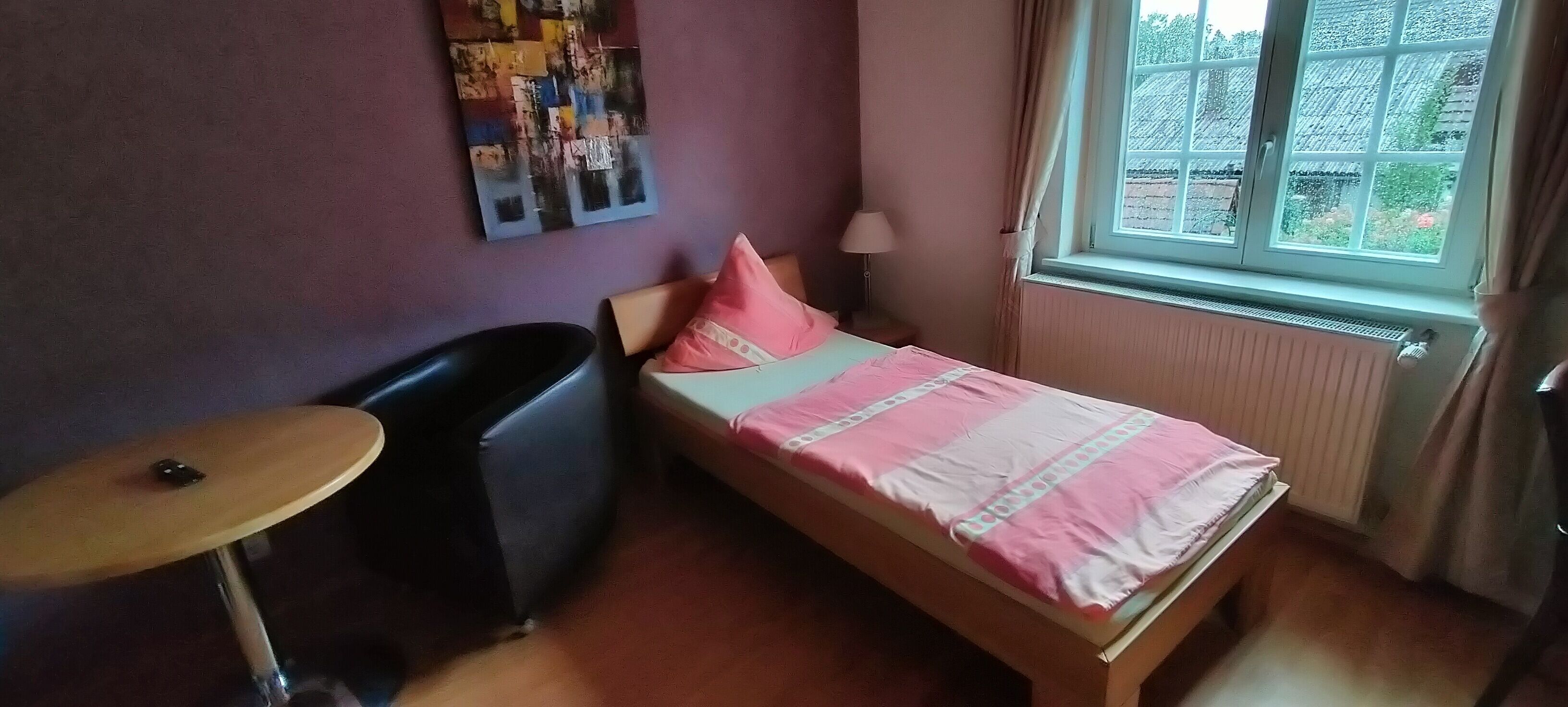 Un dormitorio con cama, una silla, una mesita, un radiador y una ventana con cortinas.